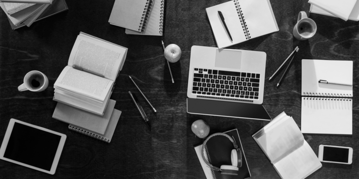 a desk crowded with notebooks, pens, laptop, tablet, phone and mugs